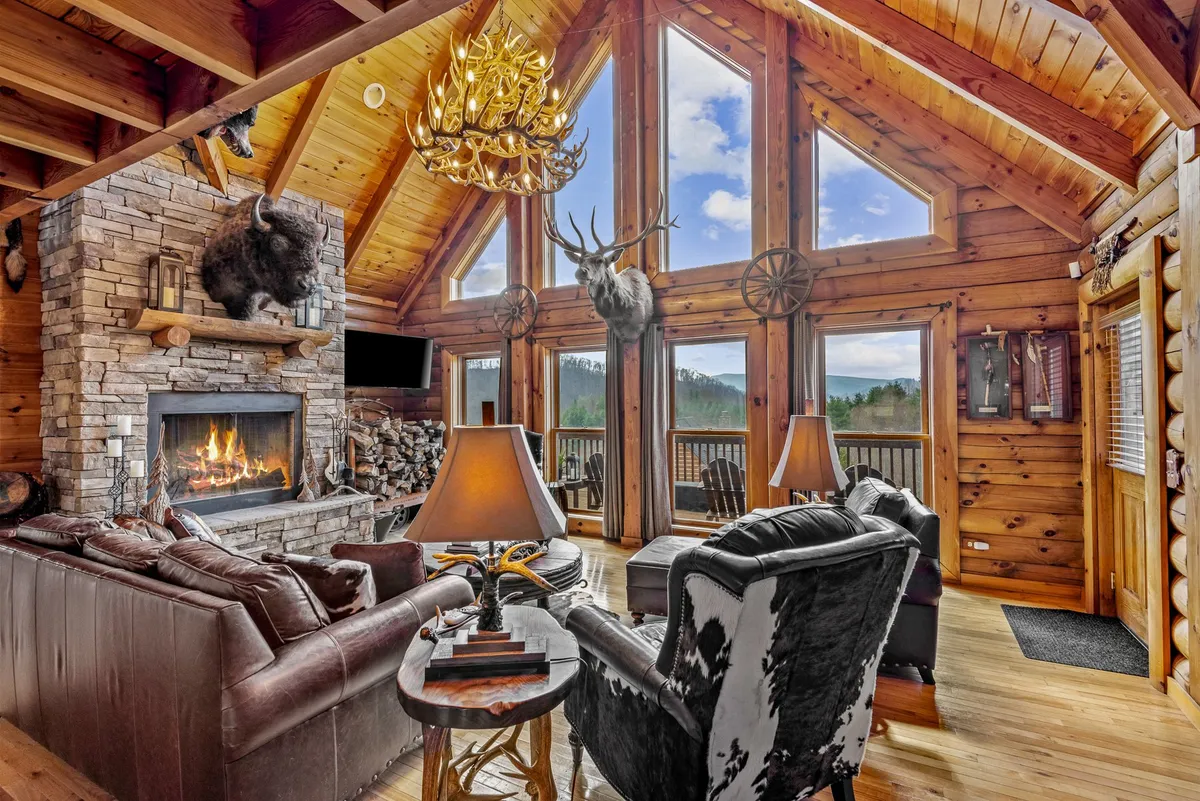 A spacious, sunlit great room featuring a dramatic floor-to-ceiling stone fireplace, large windows with expansive mountain views, an antler chandelier, exposed wooden beams, and plush seating areas. The light pouring in emphasizes the airy, warm atmosphere and the connection to the outdoors.