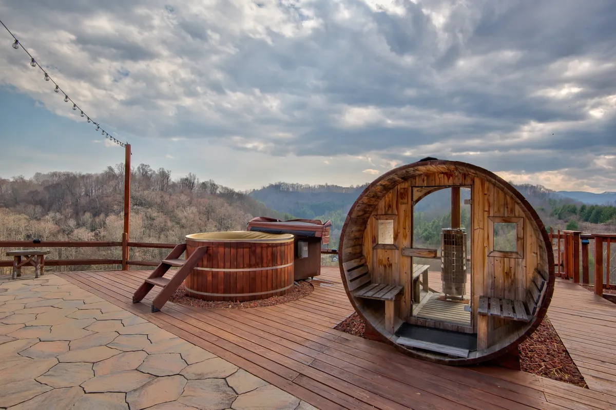 A tranquil wellness patio featuring a wooden sauna, hot tub, expansive decking, string lights, and breathtaking mountain views. The space is designed for relaxation, with options for soaking under the stars or enjoying the sauna after a long day.