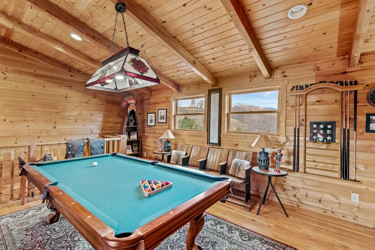 A pool table takes center stage in this inviting room, illuminated by a stained glass light fixture. Wooden benches provide ample seating for spectators or players, while decorative wall art adds character to the space. Large windows (though not the primary focus) hint at potential scenic views.