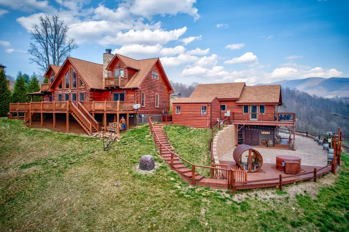 A wide view of the property, featuring log cabins, a spacious deck area, a landscaped yard, and a panoramic mountain backdrop. The setting is ideal for gatherings, barbecues, and sunset relaxation, with a hot tub visible, reinforcing the link between outdoor fun and at-home luxury.