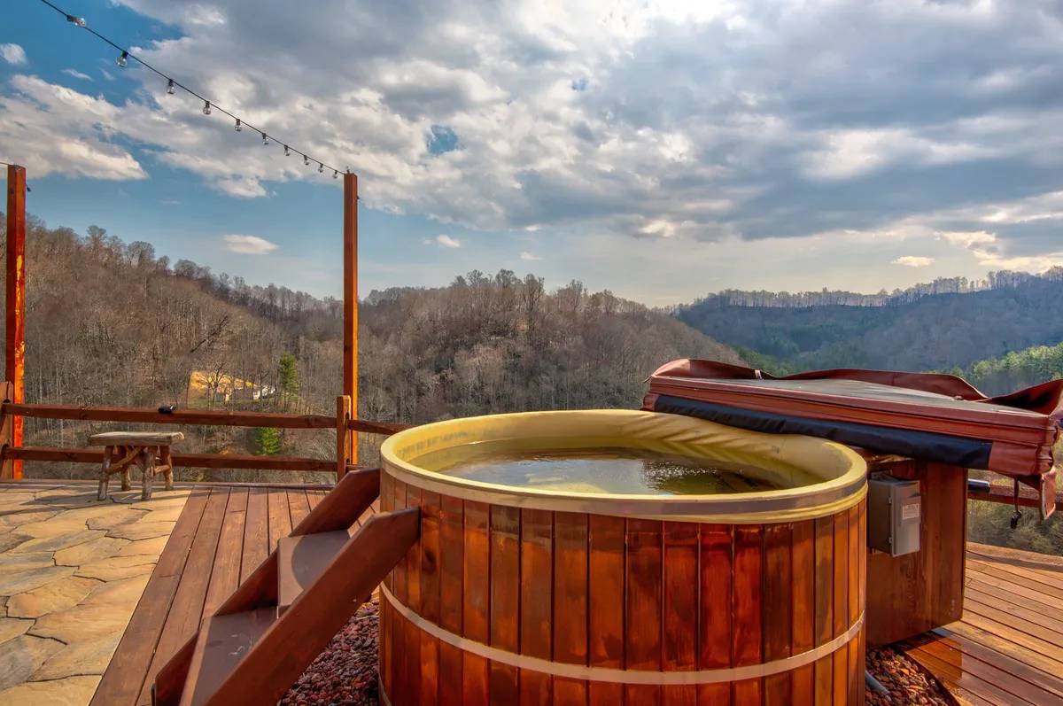 The image captures a spacious wooden deck featuring a bubbling hot tub under string lights, with expansive views of the surrounding landscape. The inviting scene suggests relaxation after a day of outdoor adventure, and the natural setting highlights the retreat’s connection to the outdoors.