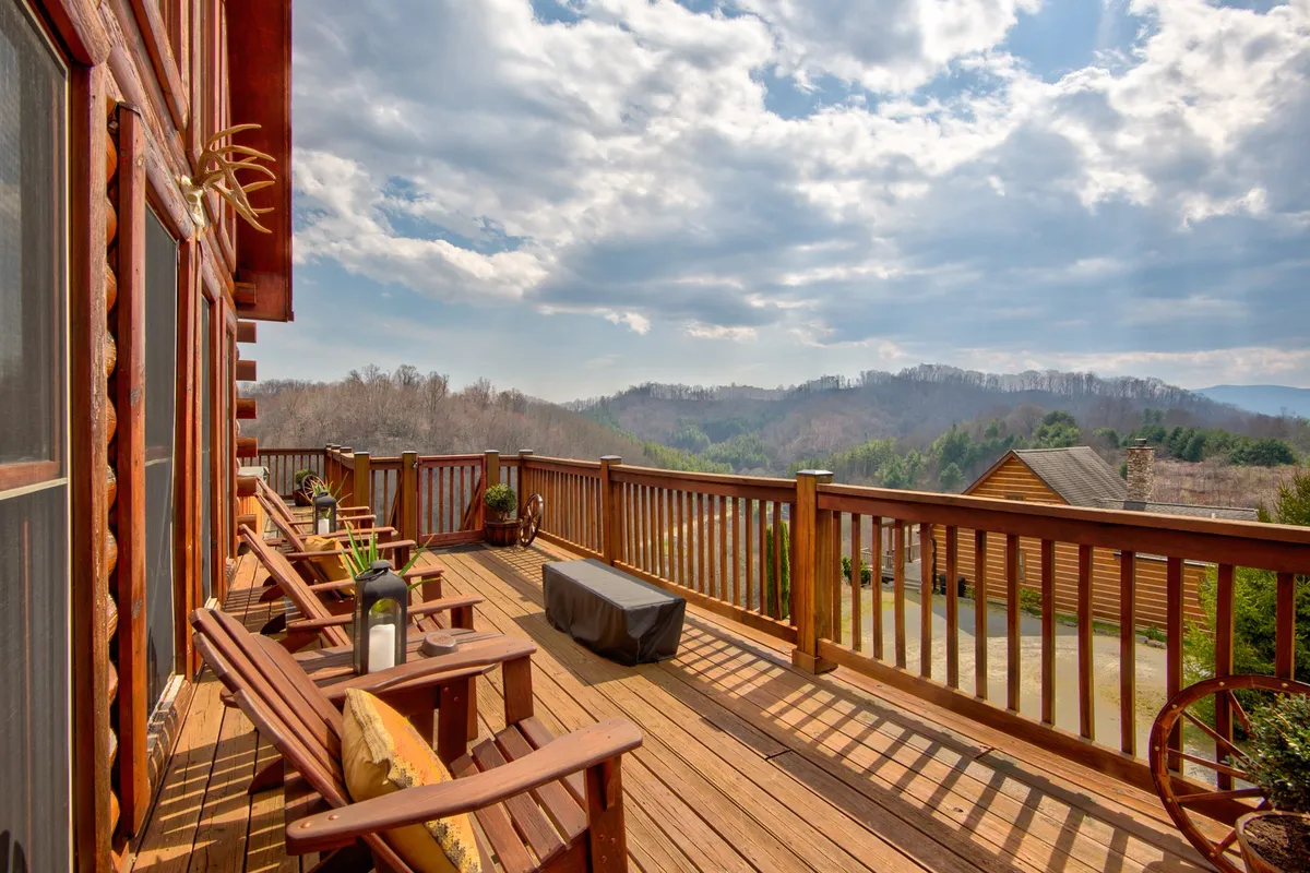 A spacious wooden deck furnished with outdoor seating offers a panoramic view of the surrounding mountains. The scene radiates serenity, with crisp morning light or the gentle glow of sunrise, inviting relaxation and inspiration. The deck's railings frame the vast landscape, making it an ideal spot for both leisure and contemplation.