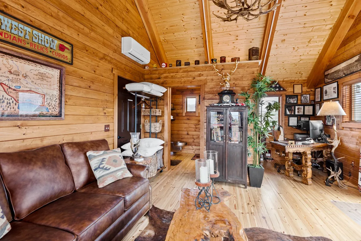 A cozy, inviting living area featuring a leather sofa, wooden table, antler chandelier, decorative wall art, and a dedicated home office area. Natural light fills the room, and the space blends comfort with functionality for both relaxation and remote work.
