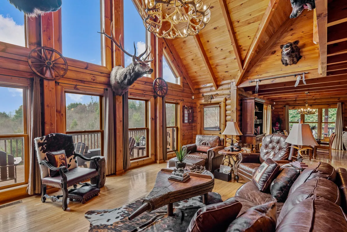 A cozy and welcoming living area with rustic furnishings, large windows offering panoramic views, a prominent chandelier, animal mounts, and rich wooden beams. The open space flows seamlessly toward adjacent areas, balancing comfort and scenic beauty.
