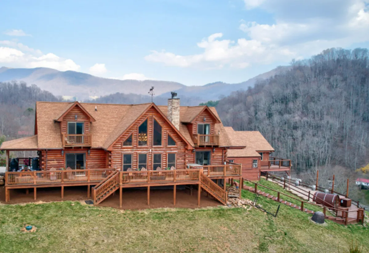 The image showcases a classic log cabin perched against a sweeping mountain backdrop, with a spacious deck offering panoramic views. The inviting structure features a prominent stone chimney, ample deck space for relaxing or dining, and is surrounded by lush greenery, emphasizing the serene and private setting.